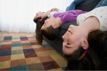 Side view of group of smiling siblings in casual clothes hanging their heads of sofa to floor while spending time together at home — Stock Photo