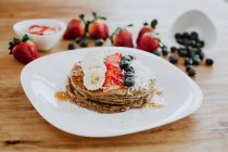Stapel leckerer Pfannkuchen auf Teller serviert mit Bananenstücken und Erdbeeren und frischen Blaubeeren mit Kokosflocken zum Frühstück — Stockfoto