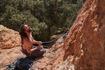 Desde arriba vista del escalador masculino sin camisa barbudo activo sentado en el borde del acantilado y sosteniendo la cuerda en las manos mientras mira hacia arriba - foto de stock