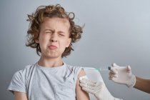 Enfermeira de colheita em luvas de látex branco segurando ombro de menino com cabelo encaracolado enquanto dá injeção de vacina com seringa na clínica moderna — Fotografia de Stock