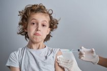Enfermera de cosecha en guantes de látex blanco que sostiene el hombro del niño con el pelo rizado mientras se administra la inyección de vacuna con jeringa en la clínica moderna - foto de stock
