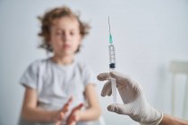 Mano de corte en guante de látex del médico anónimo que demuestra la jeringa con medicamentos de la vacuna antes de administrar la inyección al niño - foto de stock