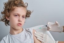 Кукурудзяна медсестра в білих латексних рукавичках тримає плече хлопчика з кучерявим волоссям, даючи ін'єкції вакцини шприцом в сучасній клініці — стокове фото