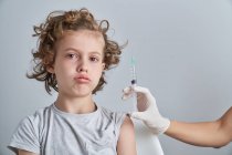 Unbekannter Arzt in Latexhandschuhen füllt Spritze mit Impfstoff aus Flasche und bereitet sich auf Injektion in die Schulter eines Jungen mit lockigem Haar vor — Stockfoto