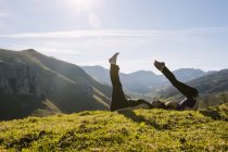 Боковой вид на мать и дочь в спортивной одежде, занимающиеся спортом с поднятыми ногами, лежа вместе на зеленой траве луга в солнечных горах в Кантабрии — стоковое фото