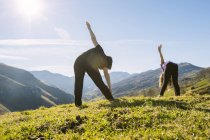 Vista lateral de madre e hija en ropa deportiva haciendo ejercicio con las piernas levantadas mientras yacen juntas sobre la hierba verde del prado en las soleadas montañas de Cantabria - foto de stock