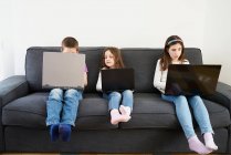 Група дітей використовує ноутбуки, сидячи на дивані вдома — стокове фото