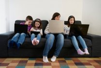Madre e hijos serios pasan tiempo juntos usando aparatos en el sofá en casa - foto de stock