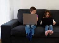 Группа детей, сидящих дома на диване с ноутбуками — стоковое фото
