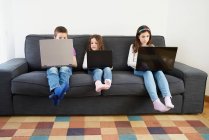 Grupo de niños que usan computadoras portátiles mientras están sentados en el sofá en casa - foto de stock
