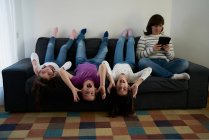 Grupo de jovens crianças alegres em roupas casuais se divertindo no sofá sentado de cabeça para baixo, enquanto sua mãe lendo livro em tablet — Fotografia de Stock