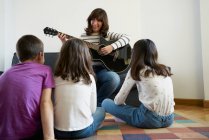Весела жінка в повсякденному одязі сидить на зручному дивані і грає на акустичній гітарі для групи дітей, сидячи на килимі на підлозі в затишній вітальні — стокове фото