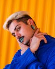 Androgynous male with blue lips and in stylish wear standing on background of yellow wall in city while looking at camera and touching face — Stock Photo