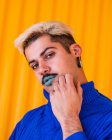Androgyner Mann mit blauen Lippen und in stylischer Kleidung, der vor dem Hintergrund der gelben Mauer in der Stadt steht, während er in die Kamera schaut und das Gesicht berührt — Stockfoto