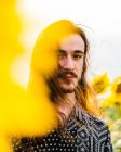Heiterer, emotionsloser Hipster-Rüde mit langen Haaren steht in gelbem Sonnenblumenfeld und blickt in die Kamera — Stockfoto