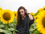 Serene unemotional hipster male with long hair standing in yellow sunflower field and looking at camera — Stock Photo