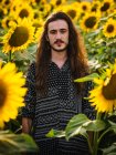 Sérénissime hipster mâle aux cheveux longs debout dans le champ de tournesol jaune et regardant la caméra — Photo de stock