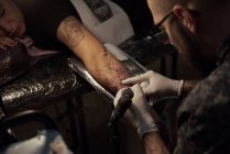 Dall'alto del tatuaggio maschile irriconoscibile che utilizza la macchina e fa il tatuaggio sul braccio del cliente del raccolto nel salone — Foto stock