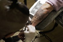 Dall'alto del tatuaggio maschile irriconoscibile che utilizza la macchina e fa il tatuaggio sul braccio del cliente del raccolto nel salone — Foto stock