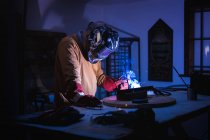 Неузнаваемый мужчина в защитных перчатках и шлеме с помощью сварочной машины во время работы в темной мастерской — стоковое фото