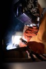 Unrecognizable male employee in protective gloves and helmet using welding machine while working in dark workshop — Stock Photo