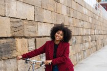 Вид спереди позитивной черной женщины, идущей с велосипедом по улице вдоль старой каменной стены и смотрящей в камеру — стоковое фото