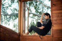 Vista lateral de descalzo solitario joven guapo étnico modelo masculino vistiendo ropa de verano hipster elegante mirando hacia otro lado mientras está sentado en el alféizar de ventanas de cabaña de madera rodeado de colchón - foto de stock