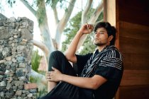 Side view of barefooted lonely young handsome ethnic male model wearing hipster stylish summer clothes looking away while sitting on wooden cabin windows sill surrounded by tress — Stock Photo