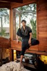 Barfuß einsame junge hübsche ethnische männliche Modell trägt Hipster stilvolle Sommerkleidung wegschauen, während sie auf Holzkabine Schlafzimmer Fensterbank von Tress umgeben sitzen — Stockfoto