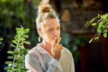 Thoughtful handsome blond male wearing hipster summer clothes looking away while standing smoking cigarette surrounded by green vegetation — Stock Photo