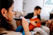 Талантливый мужчина-музыкант играет на акустической гитаре для женщины, сидящей на диване с бокалом вина и наслаждающейся песней — стоковое фото