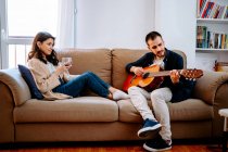 Талановитий чоловічий музикант грає на акустичній гітарі для жінки, сидячи на дивані з келихом вина і насолоджуючись піснею — стокове фото