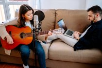 Un par de músicos sentados en el sofá y grabando canciones mientras tocan la guitarra acústica y usan el portátil - foto de stock