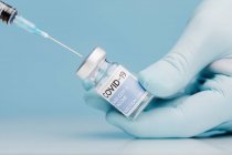 Primer plano del cultivo médico irreconocible en guantes que llenan la jeringa con la vacuna del coronavirus en un vial de vidrio sobre fondo azul - foto de stock