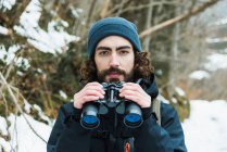 Escursionista di sesso maschile in abiti caldi in piedi in boschi invernali innevati e guardando la fotocamera in possesso di binocoli — Foto stock