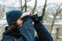 Escursionista maschio in abiti caldi in piedi in boschi invernali innevati e guardando in binocolo — Foto stock