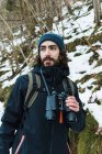 Maschio escursionista in abiti caldi in piedi in boschi invernali innevati e guardando lontano in possesso di binocoli — Foto stock