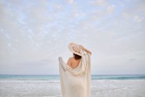 Back view female traveler in hat standing along seashore and looking away — Stock Photo