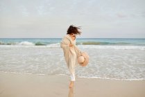 Mulher viajante encantada em roupas molhadas andando ao longo da costa no fundo de penhasco rochoso e olhando para a câmera — Fotografia de Stock