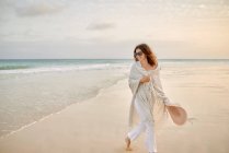 Женщина-путешественница в солнцезащитных очках и шляпе, идущая вдоль берега моря и отводящая взгляд — стоковое фото