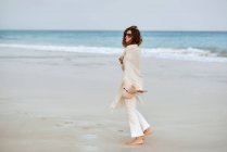Mujer viajera en gafas de sol y sombrero caminando a lo largo de la orilla del mar y mirando hacia otro lado - foto de stock