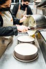 Zugeschnittene unkenntliche Köchin mit Gesichtsmaske gießt Sahne auf Kuchenform, während sie köstliches Käsekuchen-Dessert im Café zubereitet — Stockfoto