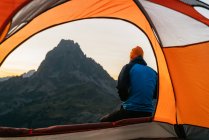 Задний вид на неузнаваемого туриста, сидящего на холме возле палатки с видом на живописные Пиренеи горного хребта утром — стоковое фото