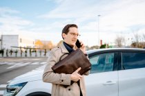Umtriebiger Unternehmer mit Attaché-Hülle steht auf Parkplatz in der Nähe des Autos und telefoniert mit dem Handy, während er das Projekt diskutiert und wegschaut — Stockfoto