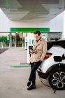 Männlicher Unternehmer in stylischem Outfit steht an Tankstelle und surft Handy, während er neben Auto mit Zapfpistole steht — Stockfoto