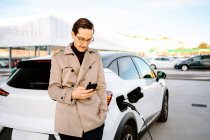 Мужчина-предприниматель в стильном наряде стоит на заправке и просматривает мобильный телефон, стоя рядом с автомобилем с топливным форсункой — стоковое фото