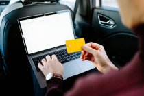 High angle of crop anonymous male entrepreneur sitting on backseat of luxury automobile with laptop and making online purchase while using plastic card — Stock Photo