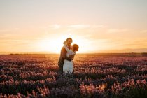 Novio feliz mirando a la cámara sonriendo mientras abraza novia de pie en el campo de lavanda en el fondo del cielo puesta del sol en el día de la boda - foto de stock