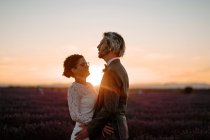 Боковой вид романтической молодоженов, стоящих лицом к лицу на просторном поле против фиолетового неба заката — стоковое фото