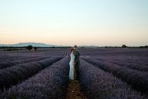 Vista lateral de pareja romántica recién casada de pie cara a cara en un campo espacioso contra el cielo púrpura atardecer mirando a la cámara - foto de stock